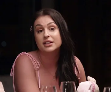 A woman with long dark hair in a pink top sits at a dinner table speaking, with glasses and a napkin in front of her.