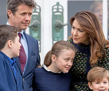 A family of five, including two adults and three children, standing closely together outdoors, smiling and interacting.