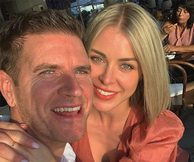 A couple smiling together, embracing at a sunlit outdoor setting, with other people in the background.