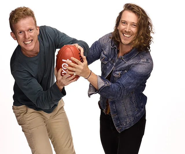 Two men smiling and holding a red football in a playful pose against a white background.