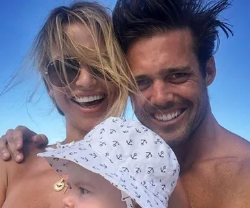 A smiling couple with a baby in a sun hat, enjoying a sunny day outdoors.