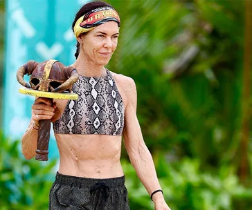 A woman wearing a snake-print top holds a tribal immunity idol on a tropical Survivor game show set.