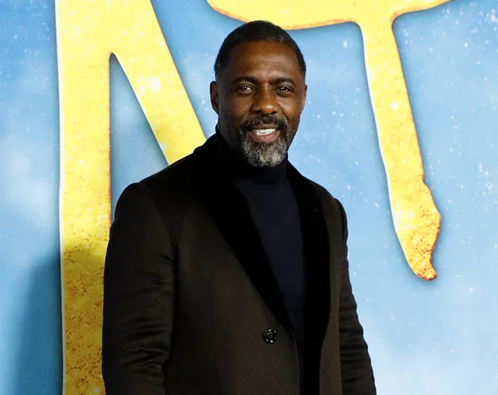Man in a dark suit stands smiling in front of a blue and yellow background.