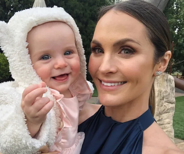 Smiling woman in a blue dress holds a cheerful baby in a fluffy white outfit outdoors.