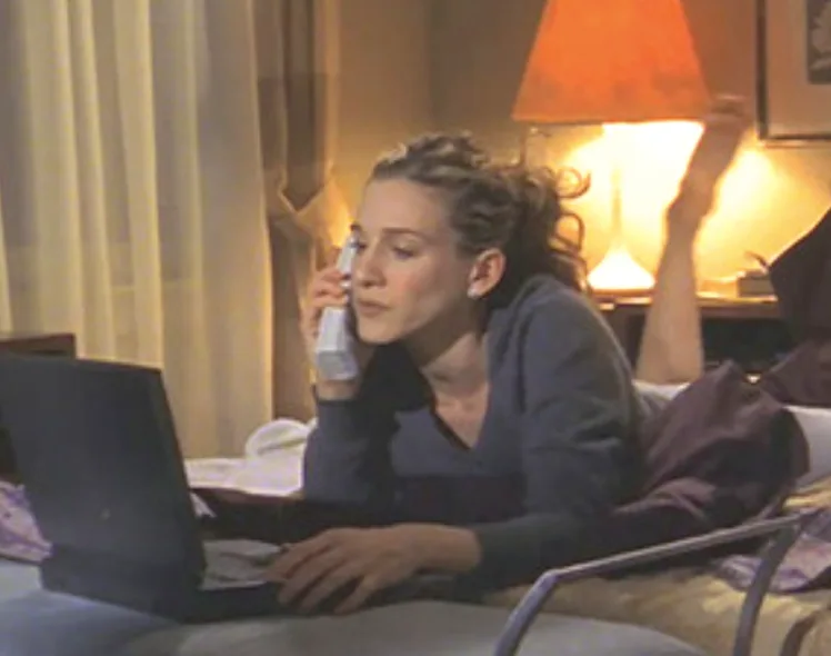 Woman lying on bed, holding phone, typing on laptop, with lamp in background.