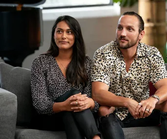 A couple sits on a couch, woman in a patterned blouse and man in a leopard print shirt, both appear attentive.