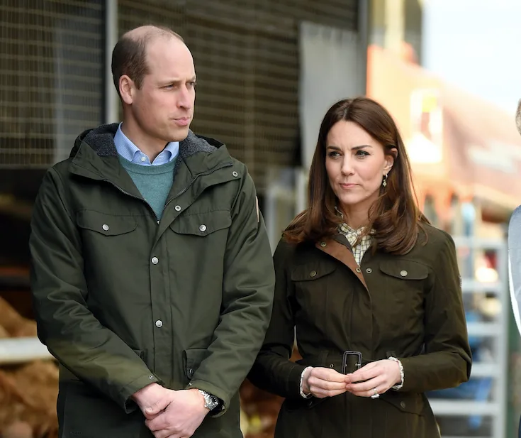 Couple standing together outdoors in matching dark coats, looking away from the camera.
