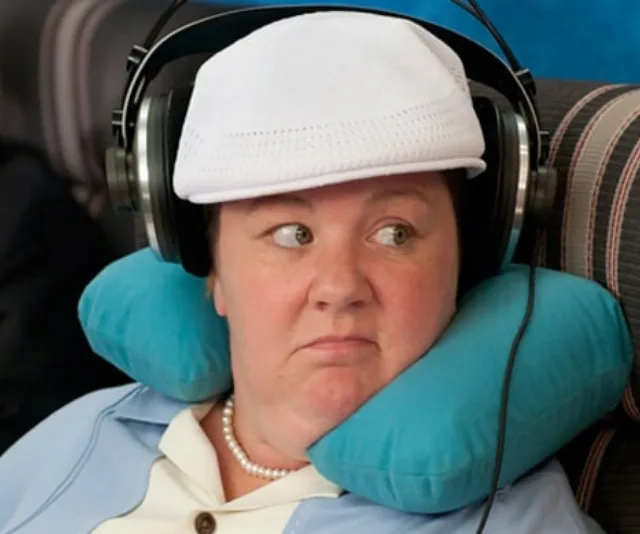 Person with headphones and a white cap, wearing a neck pillow, sitting in an airplane.