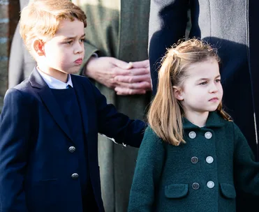 Two young children, a boy and a girl, standing outside, dressed in formal coats, looking attentively ahead.