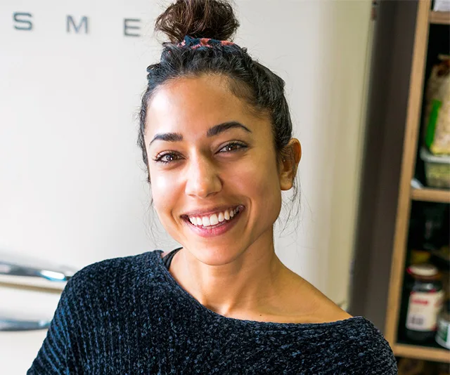 Woman with dark hair in a bun, smiling warmly, wearing a textured sweater in a kitchen setting.