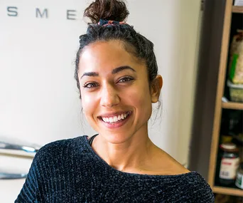 Woman with dark hair in a bun, smiling warmly, wearing a textured sweater in a kitchen setting.