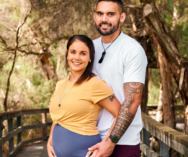 Pregnant woman and partner smiling, standing together on a forested path.