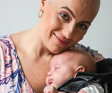 A smiling woman with short hair embraces a sleeping baby, creating a tender and warm scene.