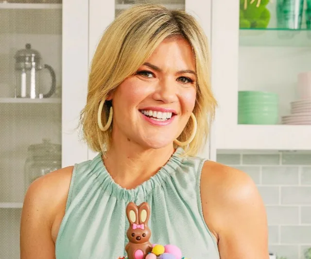 A woman in a sleeveless top holding a chocolate bunny cake and smiling in a kitchen setting.