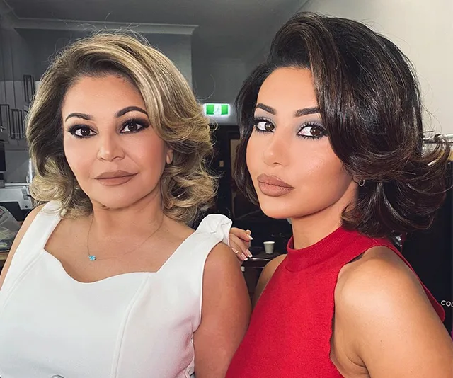 Two women with styled hair, one in a white dress and the other in a red top, posing indoors.