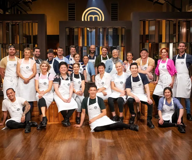 Contestants of MasterChef Australia 2020 pose together in the kitchen, wearing white aprons with the show logo in background.