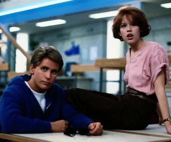 Two teenagers in a library, one sitting on a table, both looking surprised or concerned.
