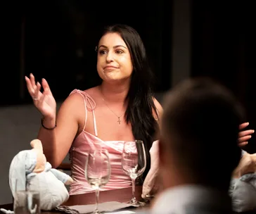 Woman in a pink dress gesturing while sitting at a table with empty wine glasses during a conversation.
