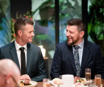 Two men in suits sitting at a dining table, smiling and conversing, with food and drinks in front of them.