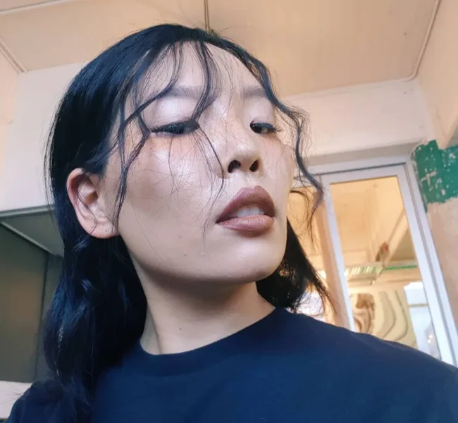 A person with long dark hair and dramatic makeup is posing confidently in an indoor setting.