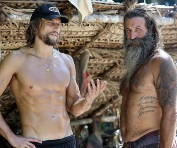 Two shirtless men with beards, one gesturing, stand by a straw hut in a rustic outdoor setting.