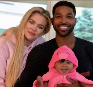 Family portrait with a woman, man, and a smiling baby in a pink animal onesie, sitting together indoors.