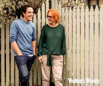 Two people smiling and talking outside near a white picket fence with greenery.