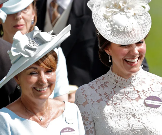 Two women in elegant hats and dresses smiling outdoors at a formal event; one in white lace, the other in light blue.
