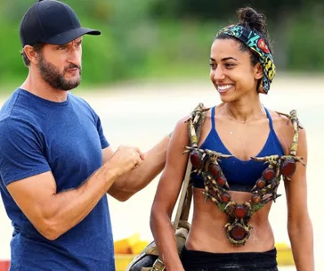 Two "Survivor" contestants, one with a necklace, are outdoors; the woman on the right is smiling.
