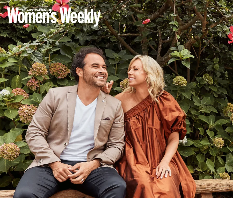 Couple sitting on a bench in a garden, smiling at each other; lush greenery in the background.