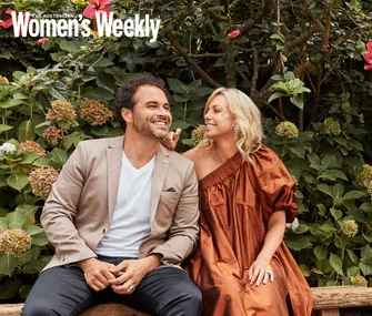 Couple sitting on a bench in a garden, smiling at each other; lush greenery in the background.