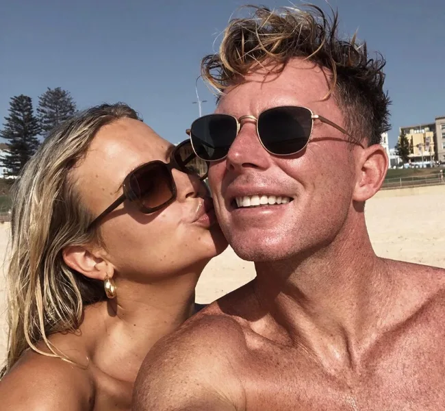 A couple smiling at the beach, wearing sunglasses, with the woman kissing the man on the cheek.