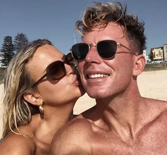 A couple smiling at the beach, wearing sunglasses, with the woman kissing the man on the cheek.