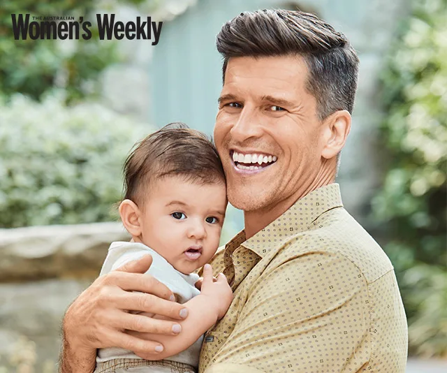 Man smiling, holding a baby, outdoors. Image from The Australian Women's Weekly.