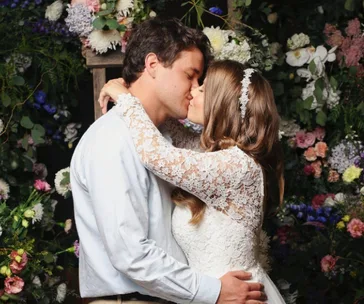 A couple kissing on their wedding day, surrounded by flowers; the bride in a lace gown and headpiece.