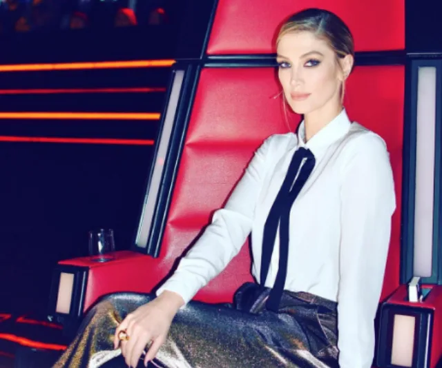 Delta Goodrem seated in a red chair on "The Voice," wearing a white shirt and black tie, with a focused expression.
