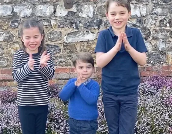 Children clapping outdoors in front of a stone wall, participating in a "Clap for Carers" event.