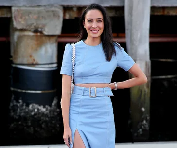 A woman in a light blue outfit stands outside on a wooden deck, smiling, with her hand on her hip.