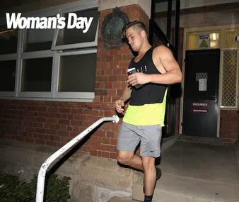 Man in athletic wear holding a phone and drink, walking down stairs outside a brick building at night. "Woman's Day" logo above.