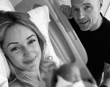 A couple smiling at the camera, one is lying in a hospital bed holding a newborn wrapped in a blanket, with a man beside them.