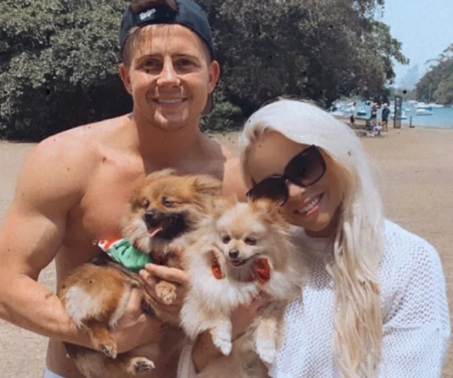 A smiling couple holding two small fluffy dogs, standing outdoors in a park.