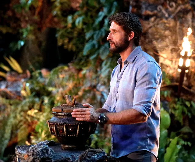 Man in casual shirt stands next to a tribal council urn in a jungle setting on a reality TV show.