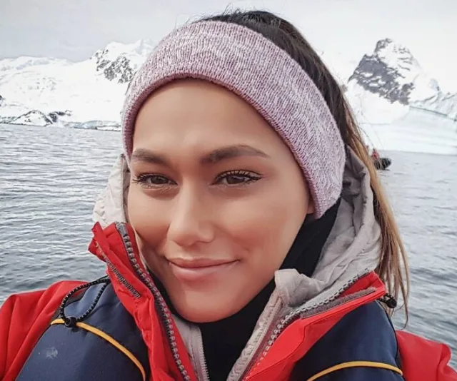 Person in red jacket and headband smiling with snowy mountains and water in the background.