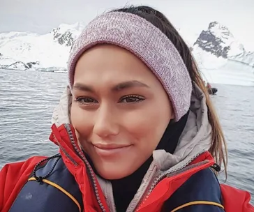 Person in red jacket and headband smiling with snowy mountains and water in the background.