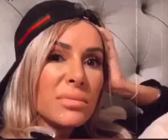 A person with long hair and a cap looks towards the camera with a concerned expression, resting their head on a hand.