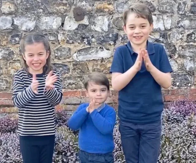Three children standing outdoors, clapping with smiles, against a stone wall background.