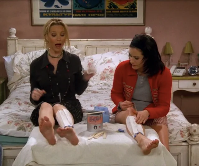Two women sitting on a bed applying wax strips to their legs, with waxing supplies between them.