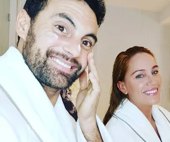 Two smiling people in white robes; one applies cream to their cheek.
