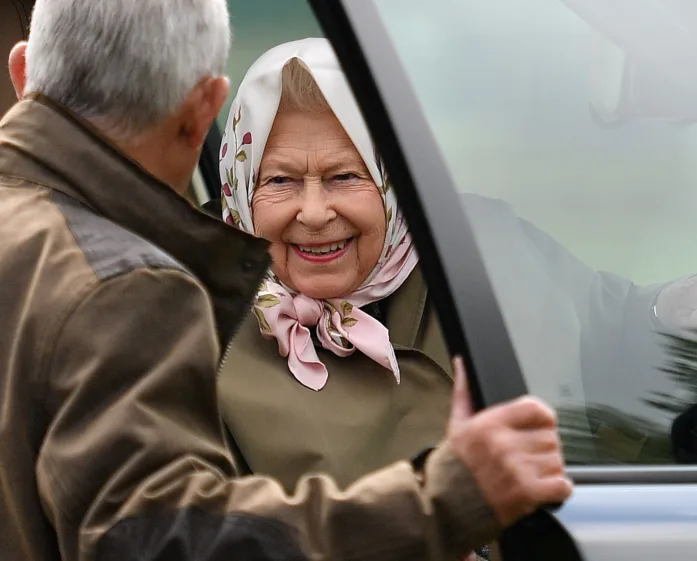 A smiling elderly woman in a headscarf is greeted by a man holding a car door open for her.