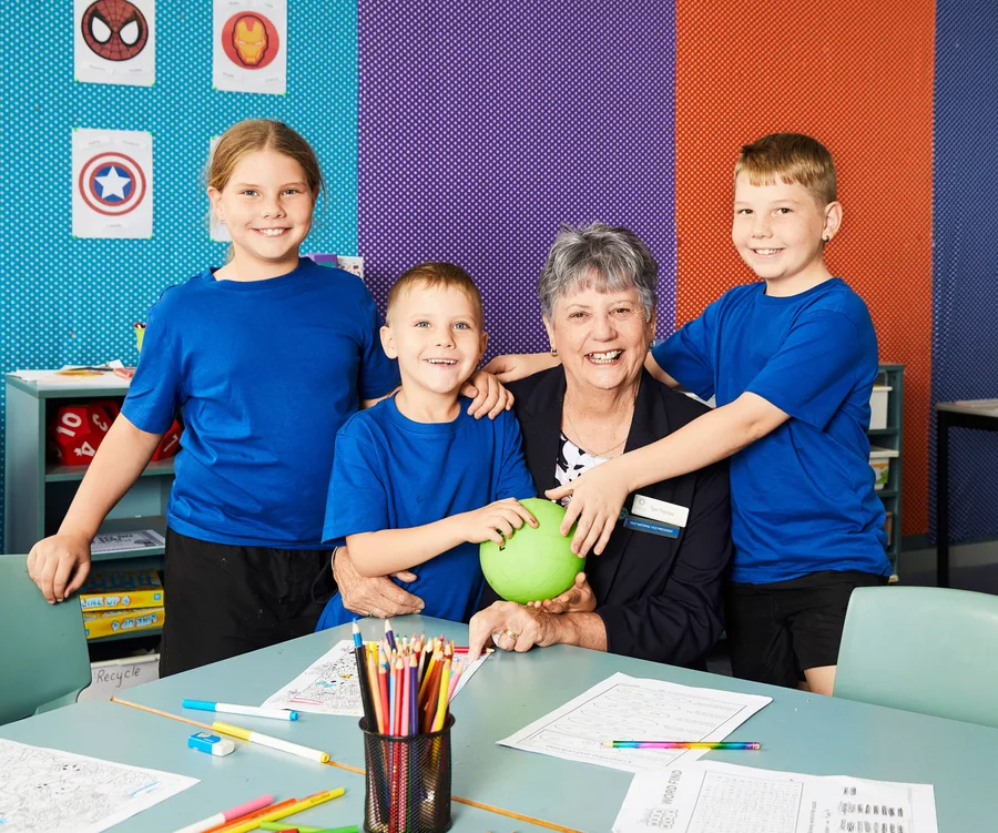 Children in blue shirts pose with a smiling elderly woman in a colorful classroom with superhero posters.